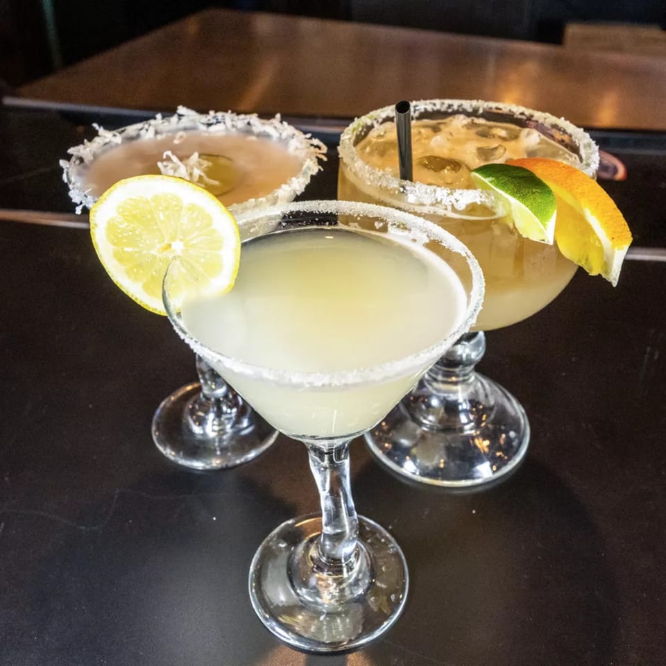 Three margaritas with salt rims and citrus garnishes on a dark bar top.