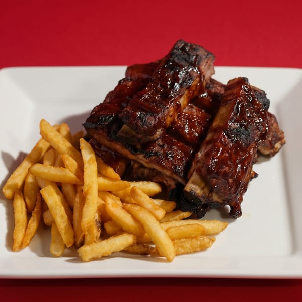 Plate of glistening BBQ ribs with a side of golden french fries against a red background.
